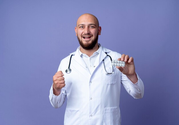 Gioioso giovane medico maschio calvo che indossa veste medica e stetoscopio tenendo le pillole e mostrando sì gesto isolato su sfondo blu