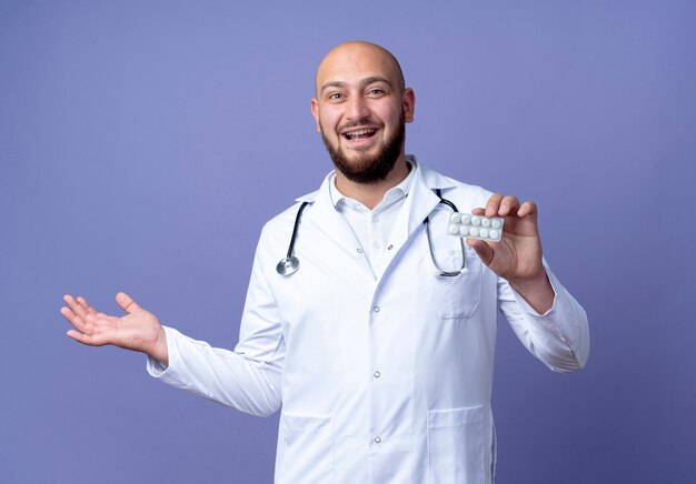 Gioioso giovane medico maschio calvo indossando abito medico e stetoscopio tenendo pillole e punti con mano a lato isolato su sfondo blu