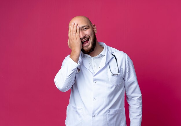 gioioso giovane medico maschio che indossa abito medico e stetoscopio coperto gli occhi con la mano