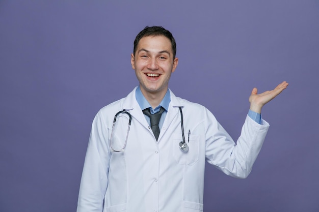 Gioioso giovane medico maschio che indossa accappatoio medico e stetoscopio intorno al collo guardando la fotocamera che mostra la mano vuota isolata su sfondo viola