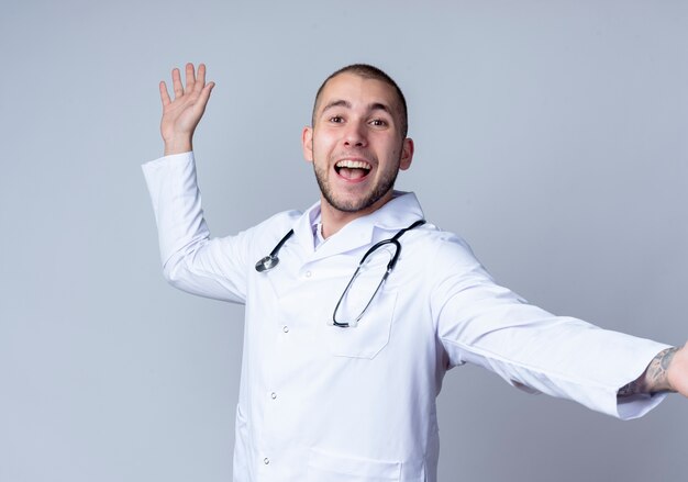 Gioioso giovane medico maschio indossa abito medico e stetoscopio intorno al collo allungando il braccio e alzando la mano isolato su bianco