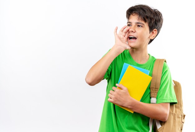 Gioioso giovane scolaro che indossa uno zaino con in mano libri che chiama qualcuno isolato sul muro bianco con spazio di copia