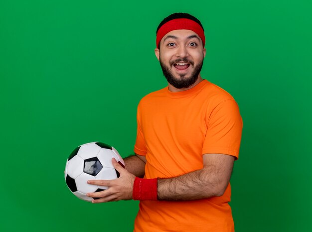 Gioioso giovane sportivo da indossare fascia e polsino tenendo fuori la palla a lato isolato su verde