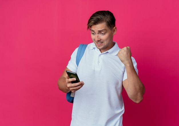 Gioioso giovane studente maschio bello che indossa la borsa posteriore che tiene e guardando il telefono e mostrando sì gesto isolato sulla parete rosa