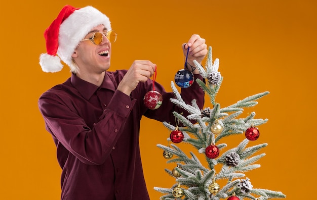 Gioioso giovane uomo biondo che indossa santa cappello e occhiali in piedi in vista di profilo vicino albero di Natale decorato su sfondo arancione