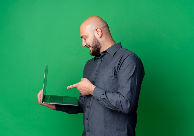 Gioioso giovane uomo calvo call center in piedi in vista di profilo tenendo e indicando il computer portatile isolato su verde