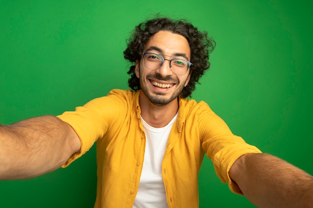 Gioioso giovane uomo caucasico bello con gli occhiali che guarda l'obbiettivo allungando le mani verso la telecamera isolata su sfondo verde