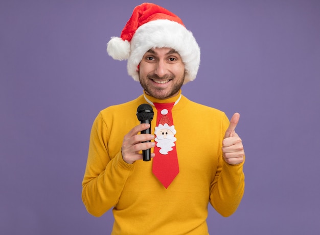 Gioioso giovane uomo caucasico indossando il cappello di Natale e cravatta tenendo il microfono guardando la telecamera che mostra il pollice in alto isolato su sfondo viola