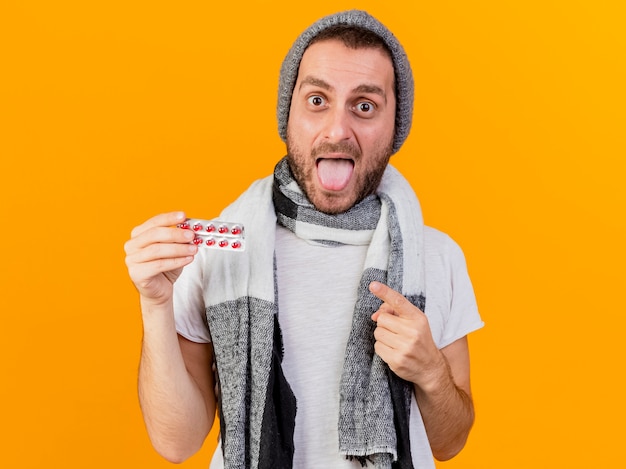 Gioioso giovane uomo malato che indossa cappello invernale e sciarpa holding e punti alle pillole che mostrano la lingua isolata su sfondo giallo