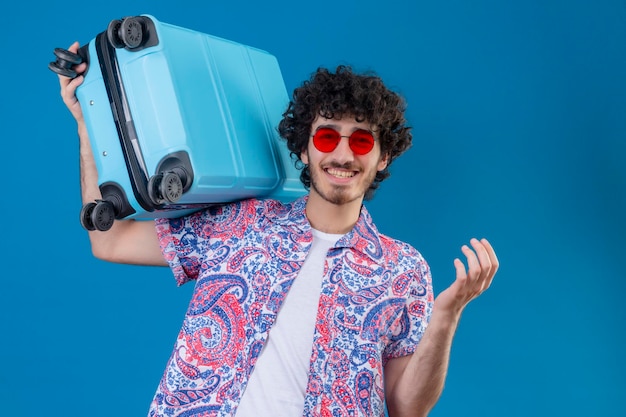 Gioioso giovane viaggiatore riccio bello uomo che indossa occhiali da sole tenendo la valigia sulla spalla su uno spazio blu isolato con spazio di copia