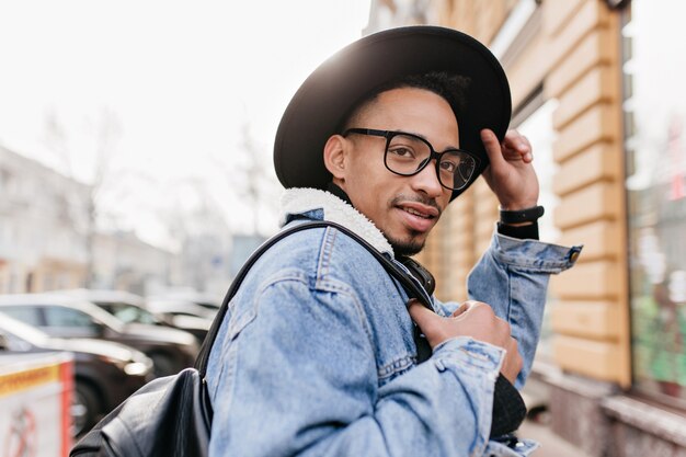 Gioioso modello maschio africano che tocca il suo cappello nero e distoglie lo sguardo sulla città. Uomo allegro del mulatto con lo zaino che cammina intorno alla città nella mattina.