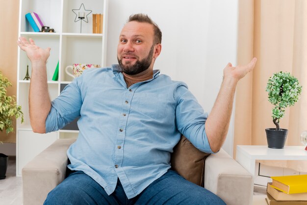 Gioioso uomo adulto slavo si siede sulla poltrona tenendo le mani aperte guardando la telecamera all'interno del soggiorno