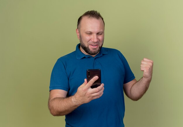 Gioioso uomo adulto slavo tenendo e guardando il telefono cellulare e facendo il gesto di sì