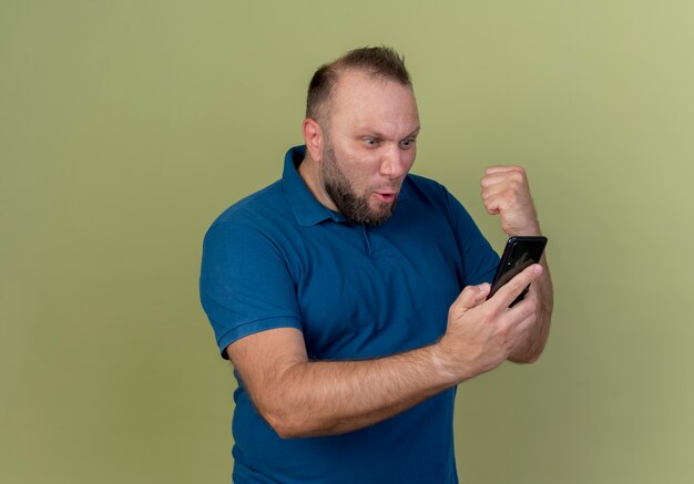 Gioioso uomo adulto slavo tenendo e guardando il telefono cellulare e facendo il gesto di sì