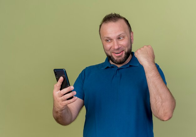 Gioioso uomo adulto slavo tenendo e guardando il telefono cellulare e facendo il gesto di sì