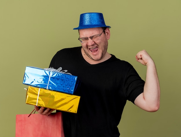 Gioioso uomo slavo adulto con occhiali ottici che indossa un cappello da festa blu tiene il pugno in mano scatole regalo e borsa per la spesa di carta paper