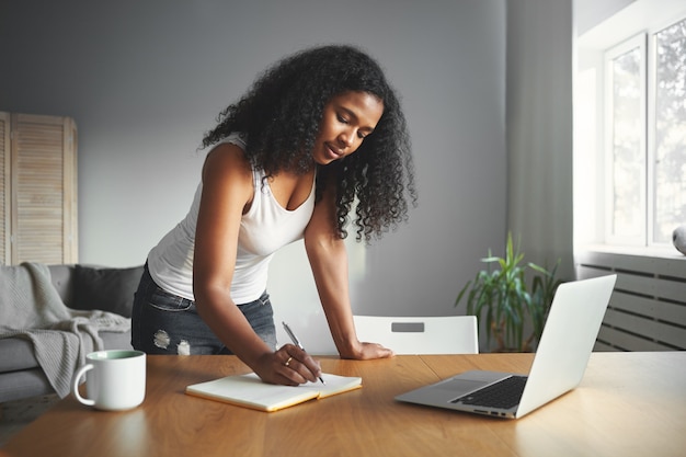 Giornata intensa della donna africana moderna che è in piedi vicino alla scrivania di legno in una stanza accogliente, annunciando qualcosa nel suo diario, avendo concentrato l'espressione facciale. Persone, stile di vita e concetto di tecnologia