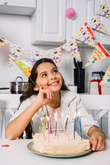 Giorno sorridente che sogna ragazza che si siede davanti alla torta di compleanno con le candele illuminate