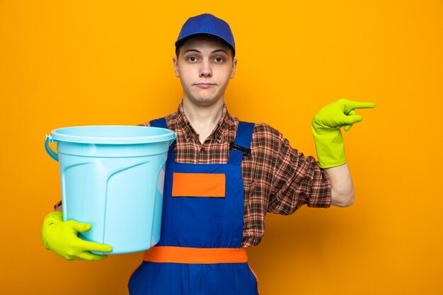 Giovane addetto alle pulizie che indossa l'uniforme e il berretto con guanti che tengono il secchio isolato sulla parete arancione con spazio di copia