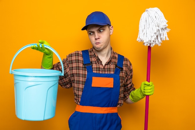 Giovane addetto alle pulizie che indossa uniforme e berretto con guanti che tengono mop e secchio isolato su parete arancione