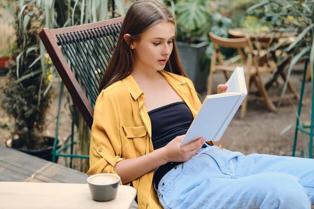 Giovane adolescente dai capelli castani attraente in camicia gialla e jeans che legge sognante un libro con caffè sulla sedia a sdraio di legno nel parco cittadino