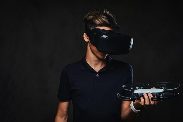 Giovane adolescente vestito con una maglietta nera indossa occhiali per realtà virtuale e tiene in mano un quadrirotore. Isolato su sfondo scuro.