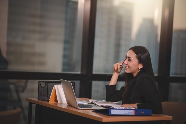 Giovane adulto asiatico intelligente donna d'affari in abito nero casual utilizzando laptop che fa gli straordinari in ufficio urbano