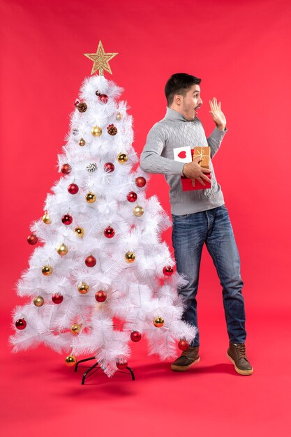 Giovane adulto bello sorpreso in piedi vicino all'albero di Natale bianco decorato e tenendo i suoi doni