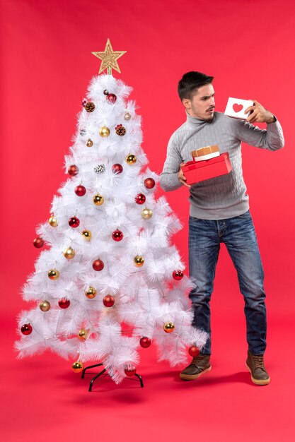Giovane adulto confuso in una camicetta grigia che sta vicino all'albero di Natale decorato e che tiene i suoi doni sul rosso