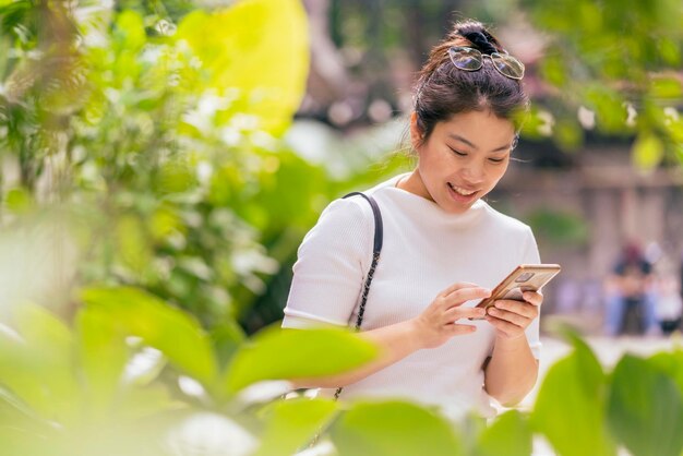 Giovane adulto femminile asiatico nomade digitale che lavora ovunque mano di comunicazione utilizzando il contatto dello smartphone e incontro con un nuovo stile di vita allegra felicità nel parco del caffè concentrato di messa a fuoco del giardino