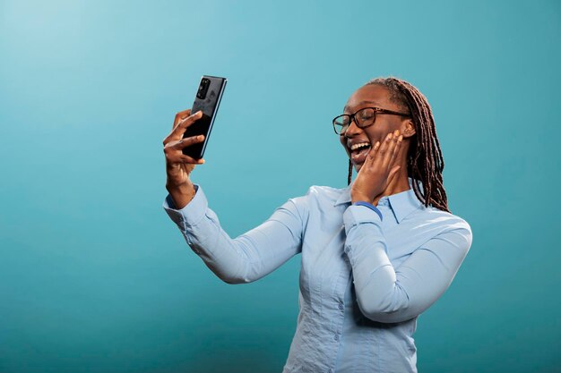 Giovane adulto soddisfatto eccitato che sorride alla fotocamera del telefono mentre scatta foto per i social media. Felice donna afroamericana sicura con il moderno smartphone touchscreen che scatta foto selfie.