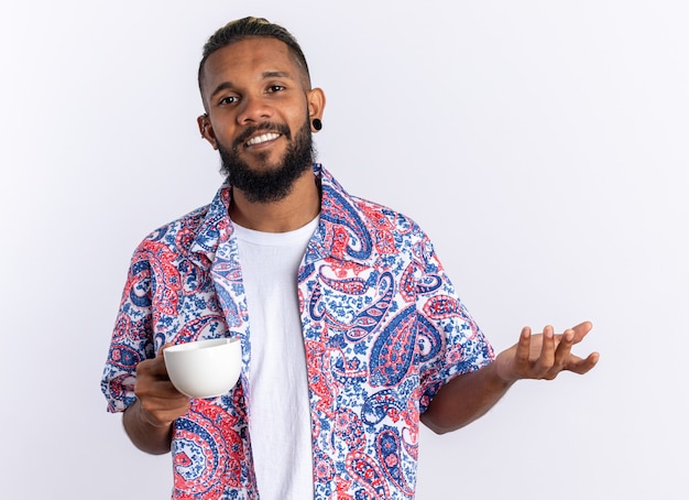 Giovane afroamericano in camicia colorata che tiene tazza felice e positivo sorridente allegramente in piedi su sfondo bianco