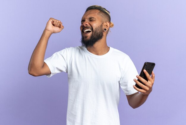 Giovane afroamericano in maglietta bianca che tiene smartphone che stringe il pugno pazzo felice ed eccitato che si rallegra del suo successo