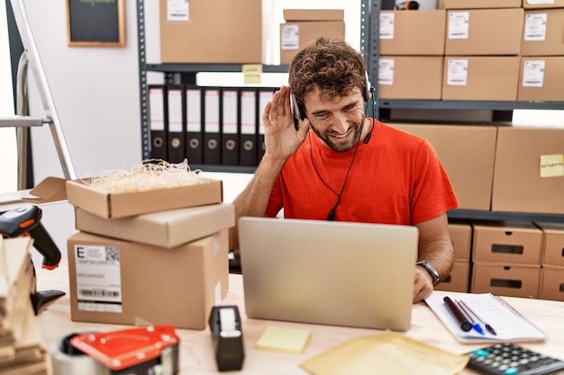 Giovane agente di call center ispanico che lavora al magazzino sorridendo con la mano sull'orecchio ascoltando un'audizione di voci o pettegolezzi. concetto di sordità.