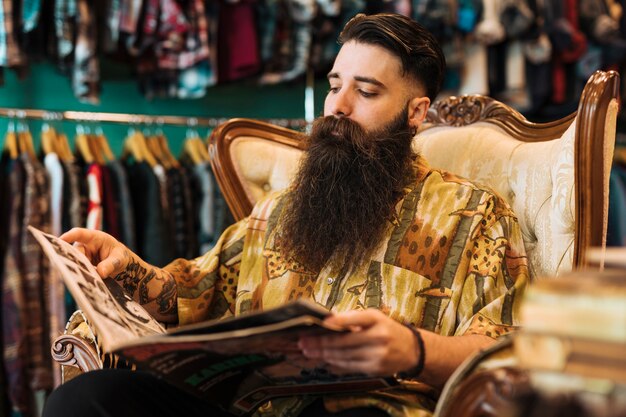 Giovane alla moda barbuto che si siede sulla sedia che esamina rivista nel negozio