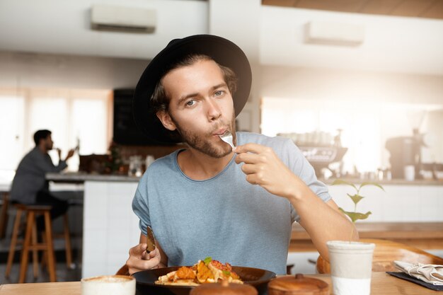 Giovane alla moda che gode dell'alimento saporito per pranzo che si siede alla tavola di legno del ristorante accogliente. Pantaloni a vita bassa affamati che portano alla moda black hat calmando la sua fame pur avendo un pasto al self-service da solo