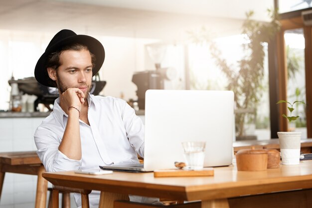 Giovane alla moda che legge e-book sul suo laptop generico, che si appoggia sul gomito e che sembra interessato. Libero professionista fiducioso che utilizza pc notebook per lavoro a distanza, godendosi il riposo durante la pausa caffè