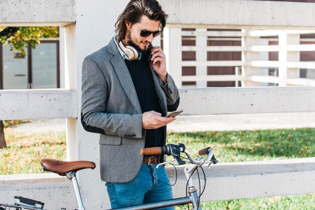 Giovane alla moda che si leva in piedi vicino alla bicicletta per mezzo del telefono mobile