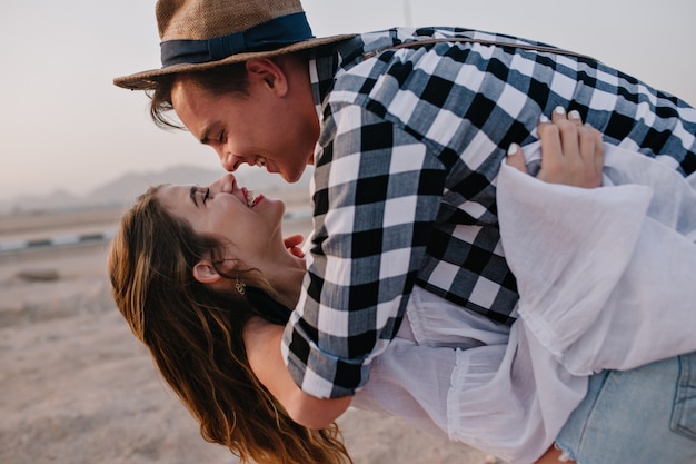 Giovane alla moda in camicia vintage e cappello elegante che balla fuori con adorabile ragazza bruna sorridente. Ritratto del primo piano delle coppie amorose che hanno divertimento sulla data all'aperto e sull'abbraccio