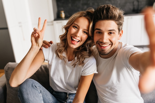 Giovane allegro che fa selfie con la sua bella ragazza a casa. Coppia con sorriso e gioia negli occhi.