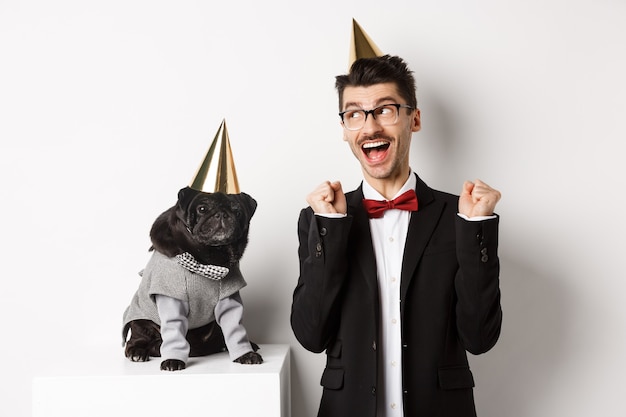 Giovane allegro che grida di gioia, cane e proprietario che indossano coni di festa di compleanno e festeggiano, ragazzo che si rallegra e fissa la macchina fotografica, sfondo bianco