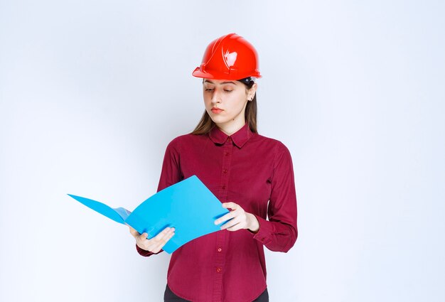 Giovane architetto femminile in casco rosso che legge note importanti su fondo bianco.