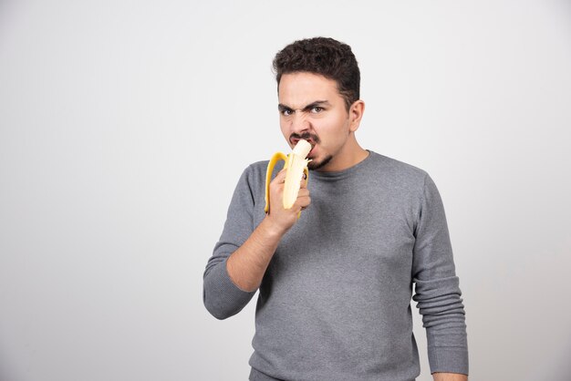 Giovane arrabbiato che mangia una banana sopra un muro bianco.