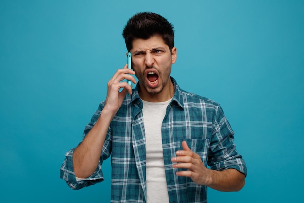 Giovane arrabbiato guardando lato parlando al telefono gridando isolato su sfondo blu