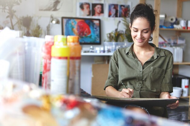 Giovane artista femminile professionale allegra di bell'aspetto che lavora al nuovo progetto creativo, disegna, fa schizzi con la matita, si sente ispirata. Concetto di persone, lavoro, occupazione, professione e hobby