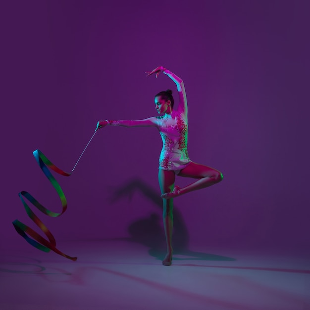 Giovane atleta femminile, artista di ginnastica ritmica danza, formazione isolato su sfondo viola studio con luce al neon. Bella ragazza che si esercita con l'attrezzatura. Grazia nelle prestazioni.