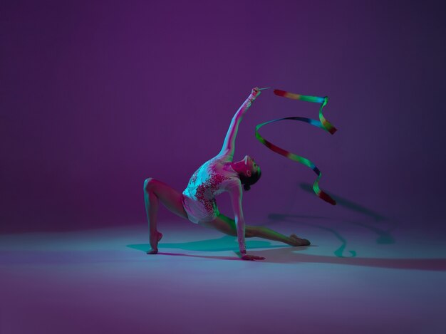 Giovane atleta femminile, artista di ginnastica ritmica danza, formazione isolato su sfondo viola studio con luce al neon. Bella ragazza che si esercita con l'attrezzatura. Grazia nelle prestazioni.