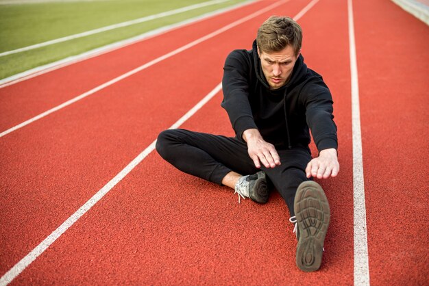 Giovane atleta maschio che si siede sulla pista di corsa che allunga la sue mano e gambe