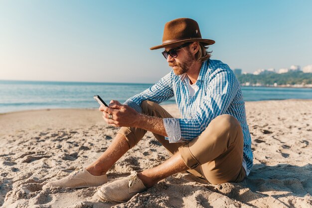 Giovane attraente hipster uomo seduto sulla spiaggia in riva al mare in vacanza estiva