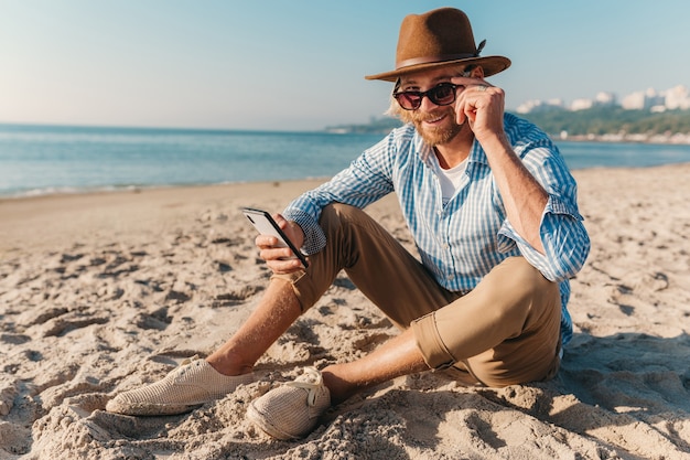 Giovane attraente hipster uomo seduto sulla spiaggia in riva al mare in vacanza estiva